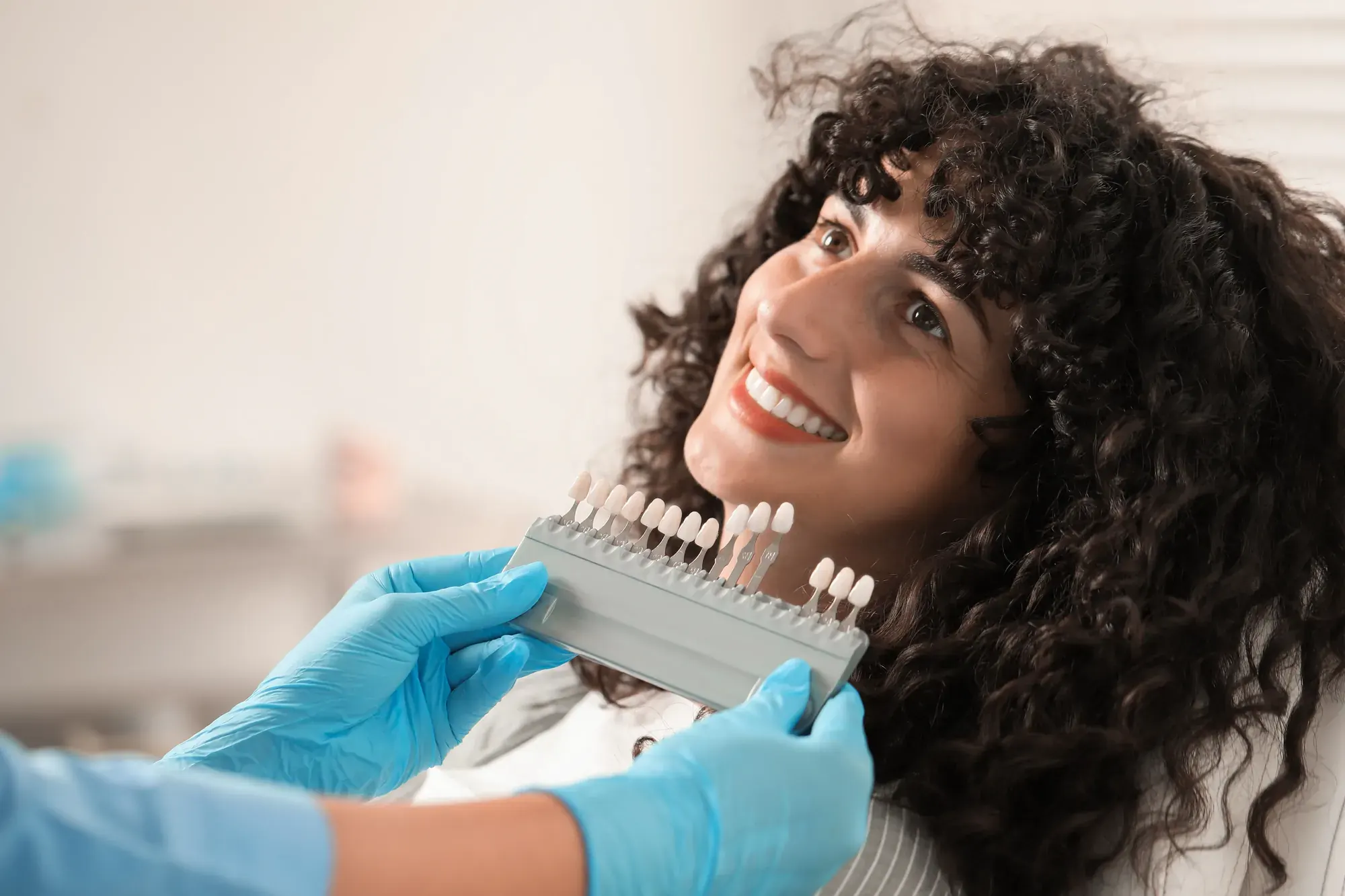 Dentist holding a porcelain shade guide to a smiling patient’s teeth to match a natural-looking crown in NYC