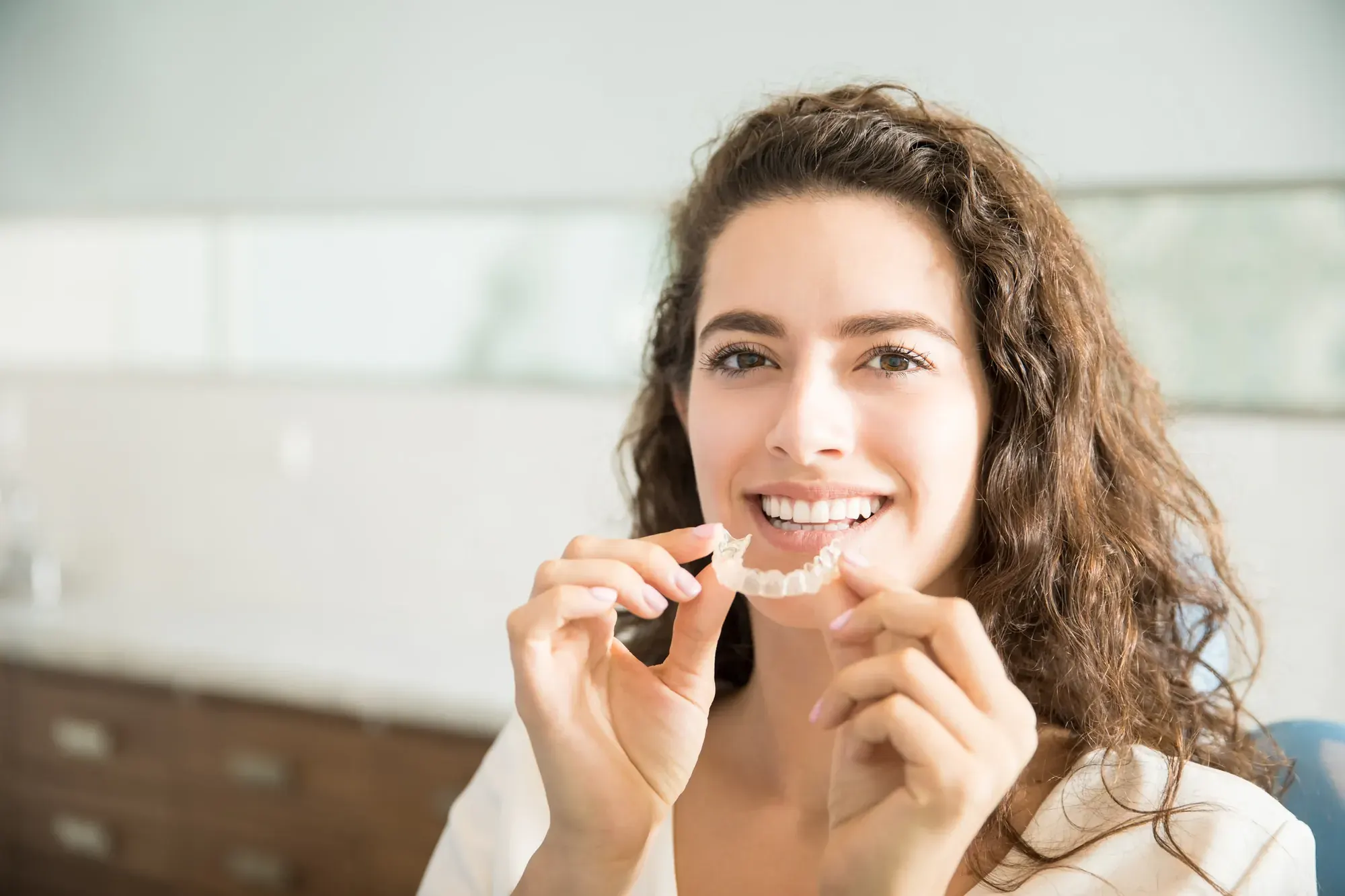 Smiling woman holding an Invisalign clear aligner next to her teeth, for Invisalign treatment in Tribeca, NYC