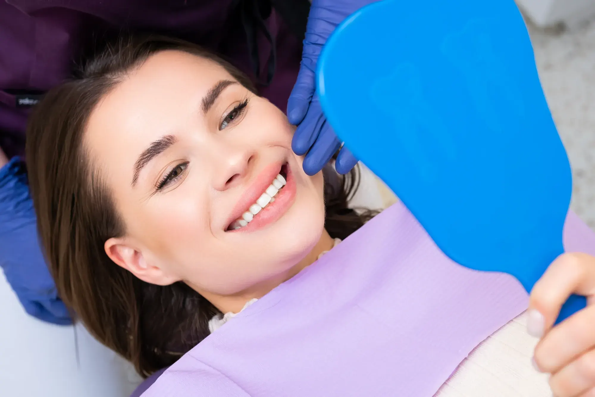 Smiling patient checking her repaired front teeth in a blue hand mirror after dental bonding in NYC