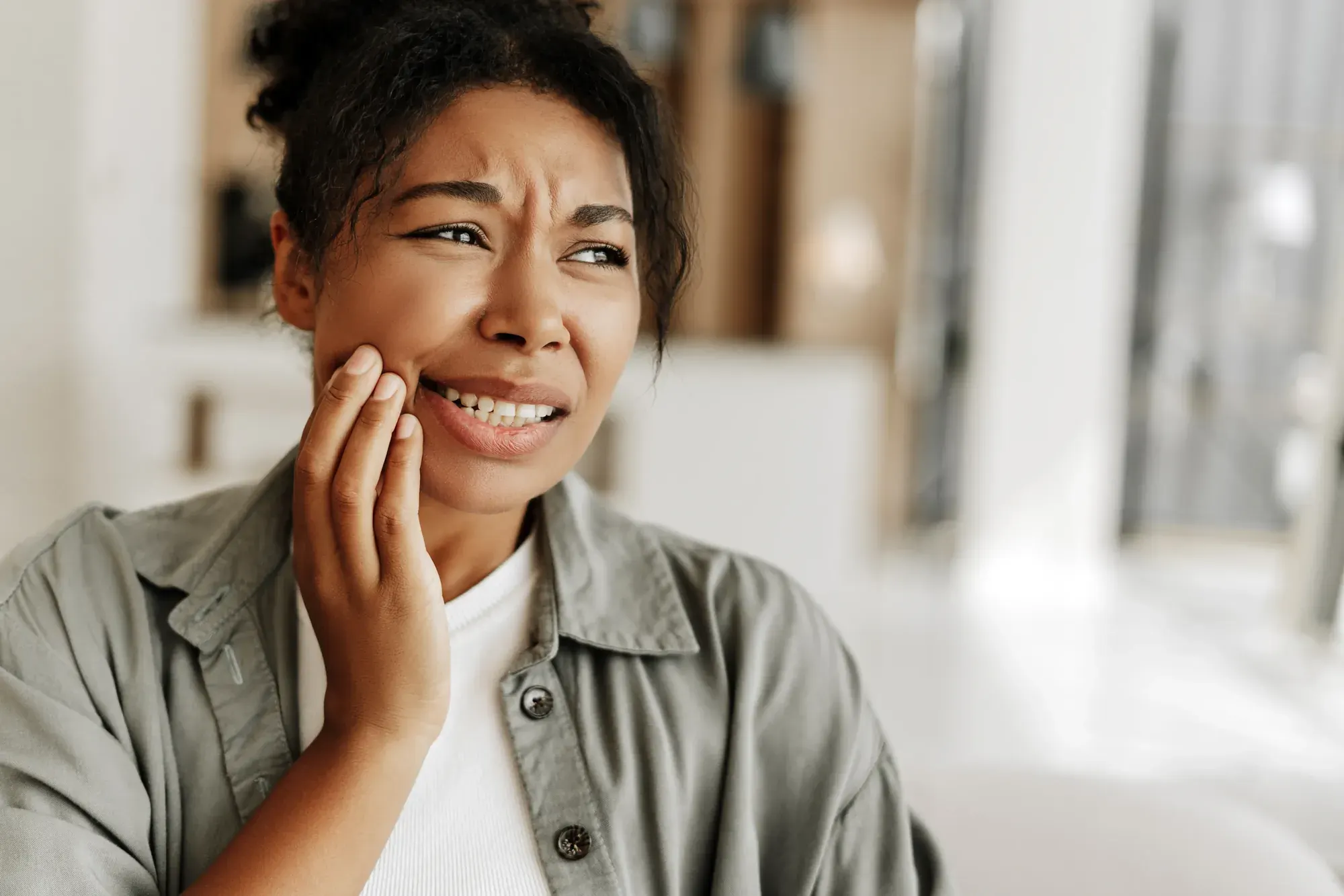 Woman at home touching her cheek with sudden tooth pain, deciding to call an emergency dentist